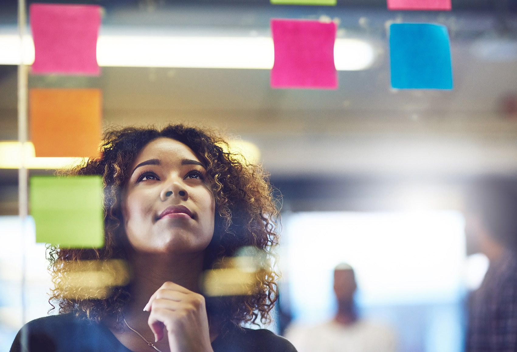 A woman thoughtfully looks at sticky notes on a glass wall, pondering ideas.